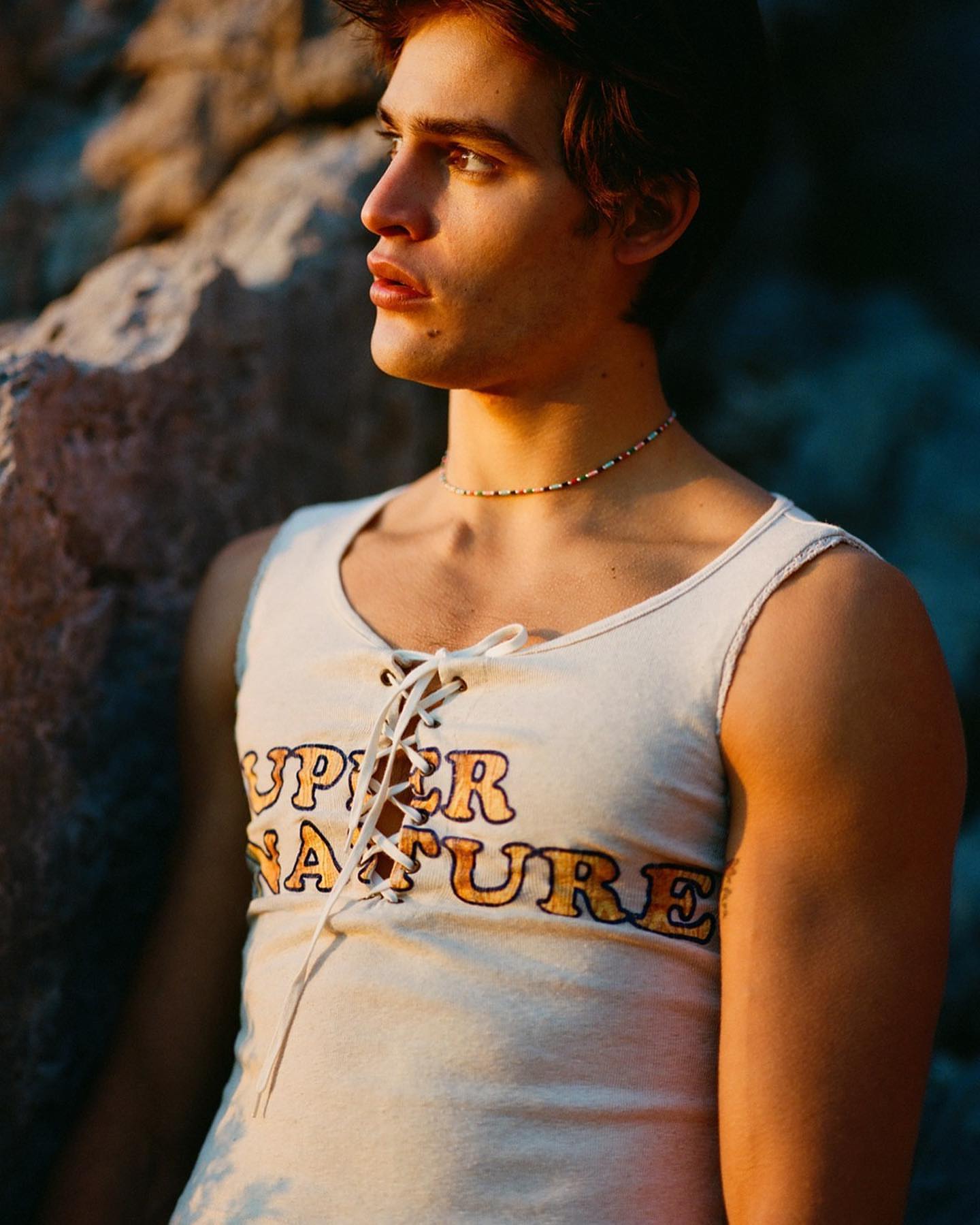 Young adult man in a lace-up tank top stands outdoors near rocks in golden light, showcasing fashion and portrait photography.