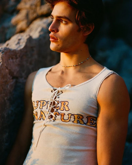Young adult man in a lace-up tank top stands outdoors near rocks in golden light, showcasing fashion and portrait photography.