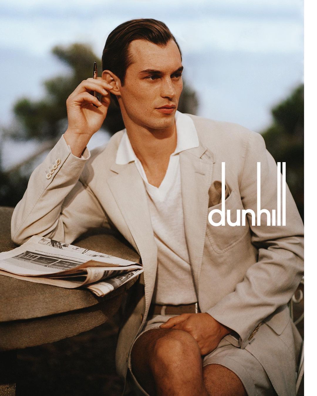 A man wearing a tan suit holds a pen up to his ear. The Dunhill logo is in the lower-right, outdoors with trees and sky in the background.