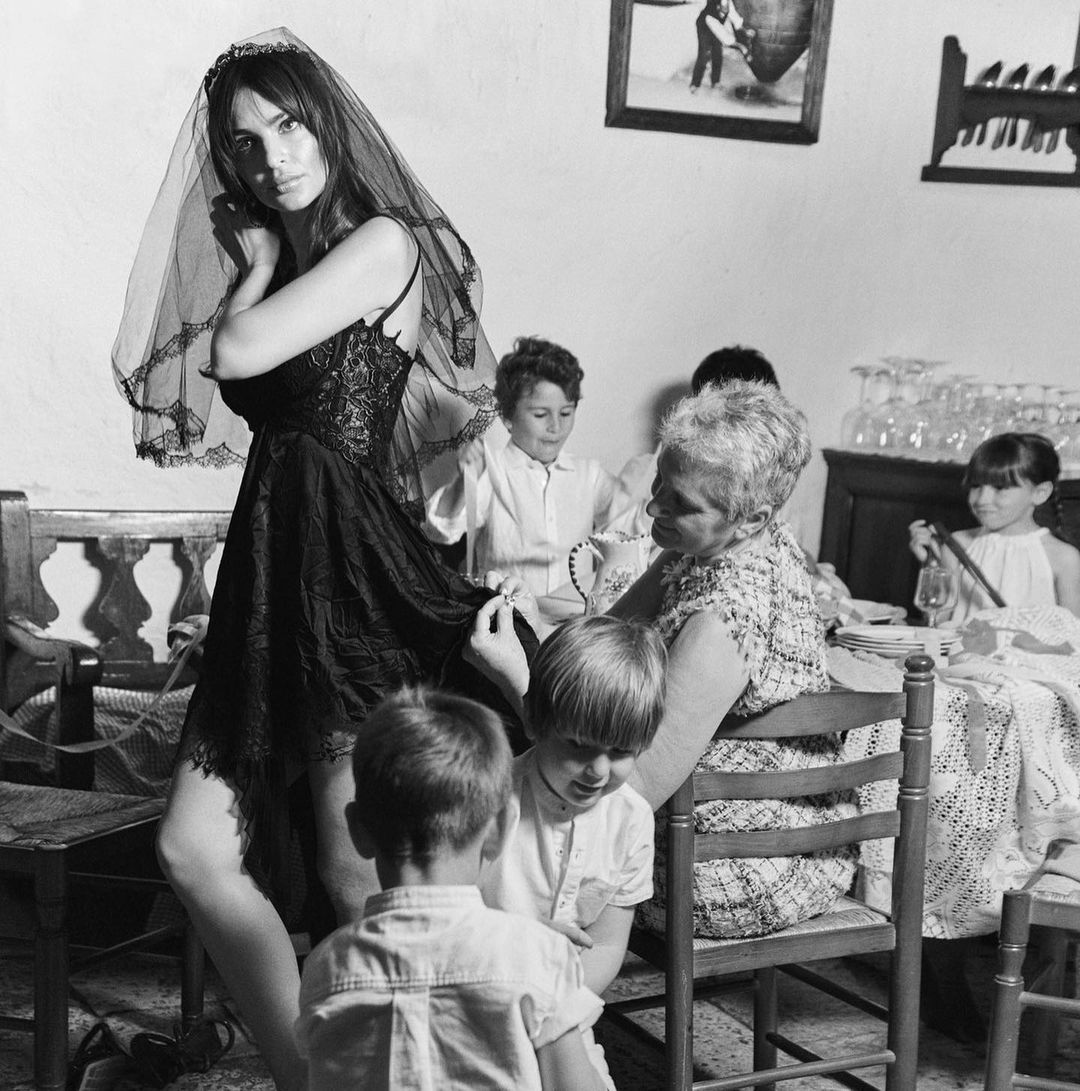 Black and white photo of a woman in a dark dress and veil standing next to a table with an older woman and children.
