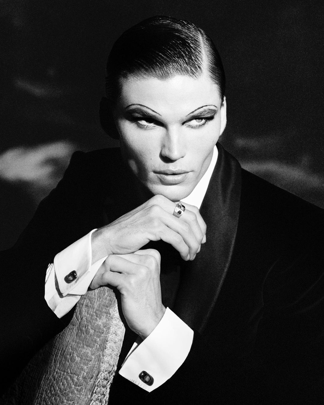 Dramatic black-and-white portrait of a model in a suit, accentuated by striking makeup and slicked-back hair, set against a moody backdrop.