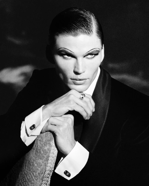 Dramatic black-and-white portrait of a model in a suit, accentuated by striking makeup and slicked-back hair, set against a moody backdrop.