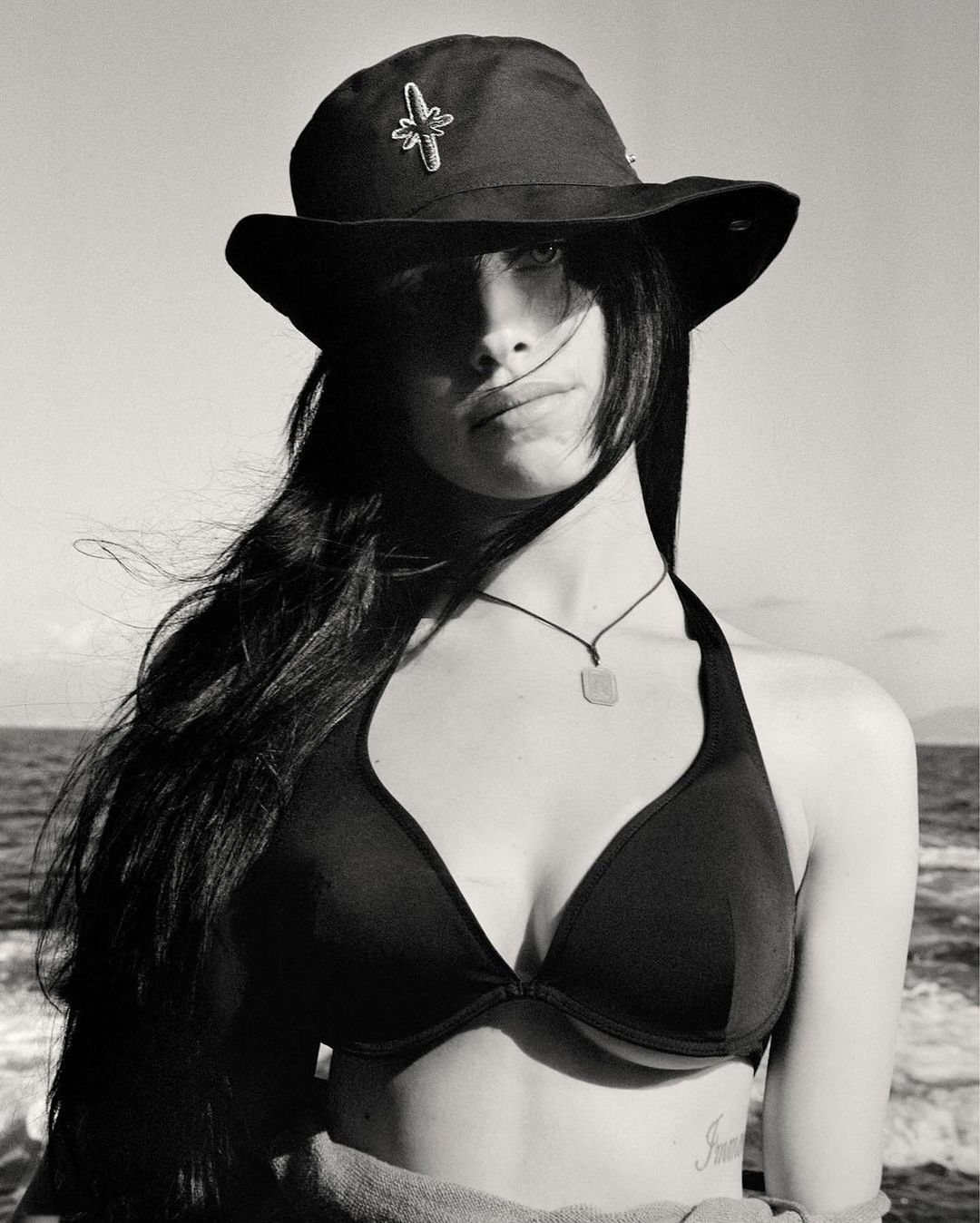 Black and white portrait of a young woman wearing a hat and bikini top on a boat, with the ocean and sky in the background.