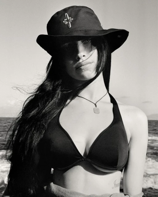Black and white portrait of a young woman wearing a hat and bikini top on a boat, with the ocean and sky in the background.