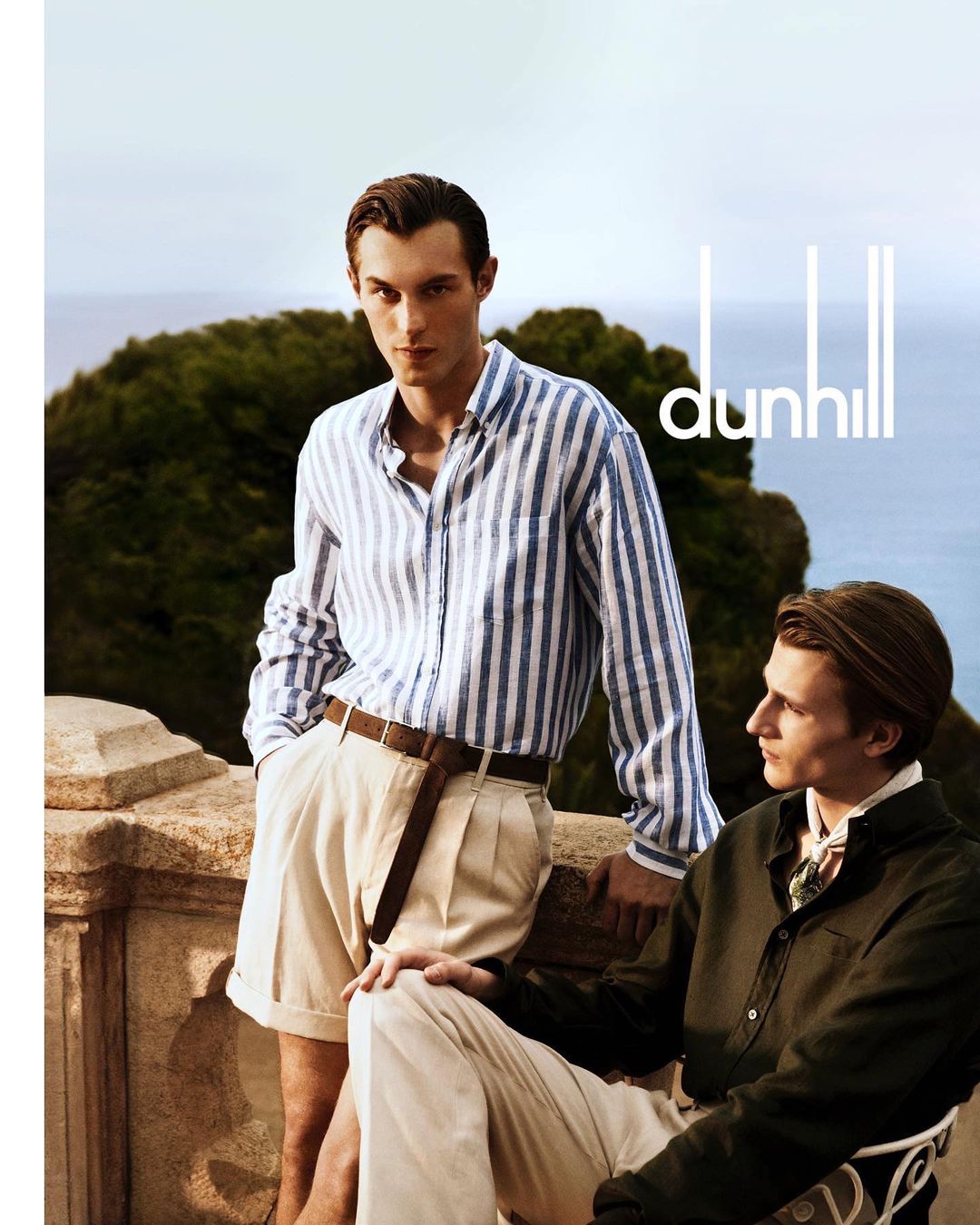 Two men pose in Dunhill fashion on a terrace with a sea view. One is standing in a blue striped shirt and the other is sitting.