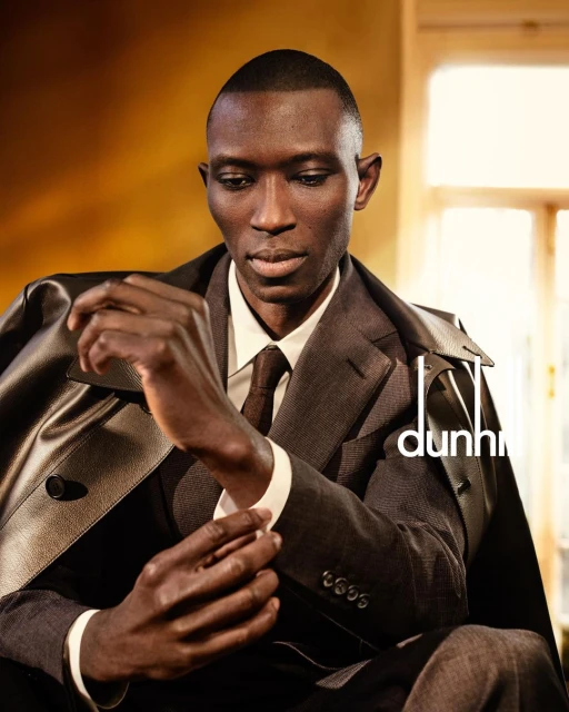 A stylish African-American male model in a grey suit adjusts his cuff indoors under soft lighting, showcasing fashion brand appeal.