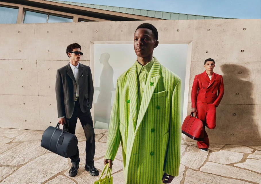Three Black male models showcase Louis Vuitton menswear in a contemporary outdoor setting, wearing tailored suits with duffel bags.