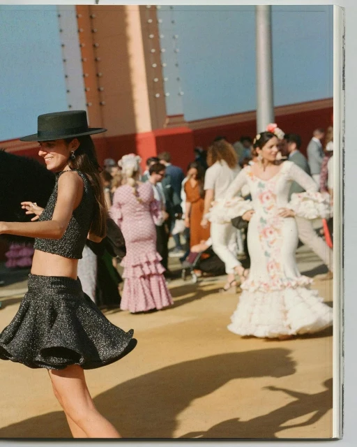 Two women in flamenco-style dresses pose in front of a crowd in an outdoor setting, with traditional architectural details visible in background.