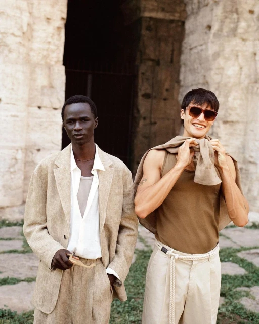 Two stylish men pose outdoors near an ancient stone wall. One wears a beige suit, the other a tank top and sunglasses.