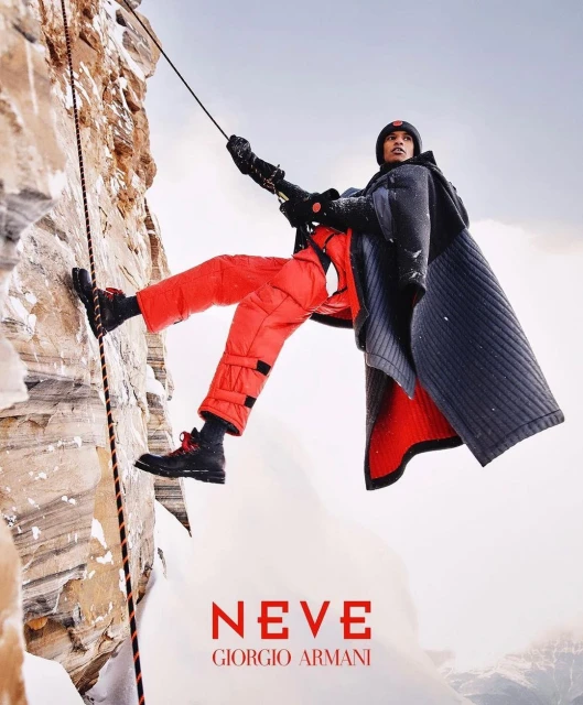 A person in red snow pants and dark coat rock climbs a mountain face with rope, set against a snowy, overcast sky, fashion advertisement.