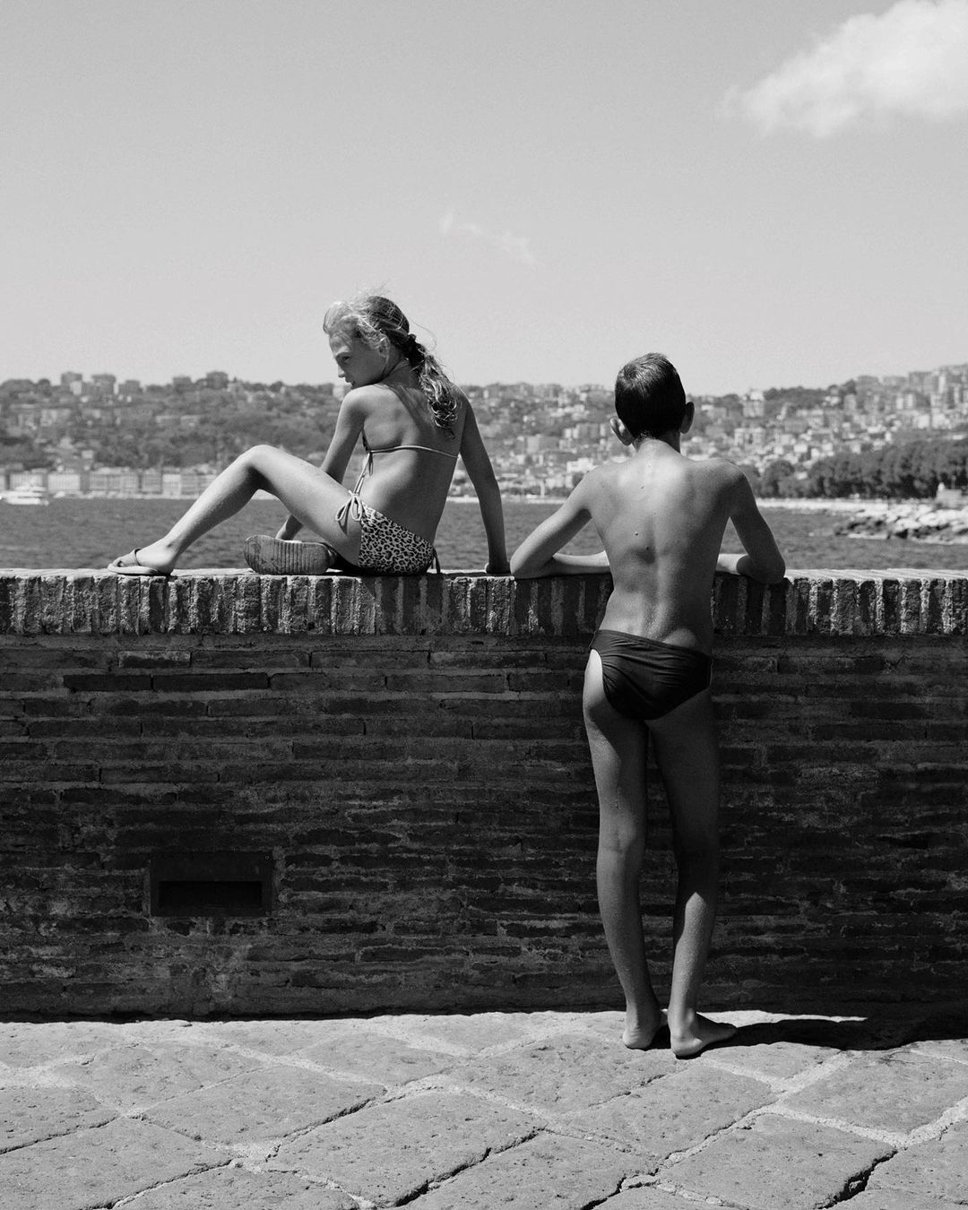 Monochrome shot of two children in swimwear on a brick wall with a cityscape and body of water in the background on a sunny day.
