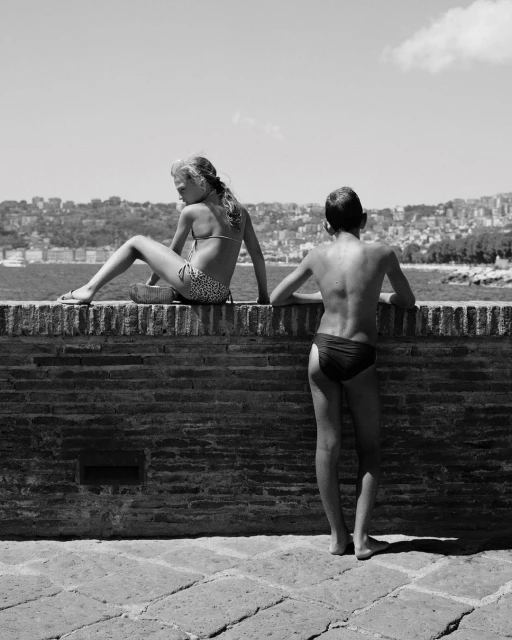 Monochrome shot of two children in swimwear on a brick wall with a cityscape and body of water in the background on a sunny day.