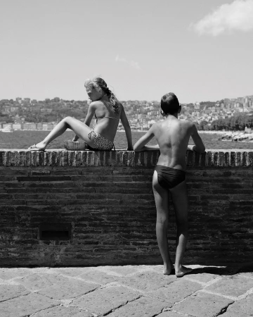 Monochrome shot of two children in swimwear on a brick wall with a cityscape and body of water in the background on a sunny day.