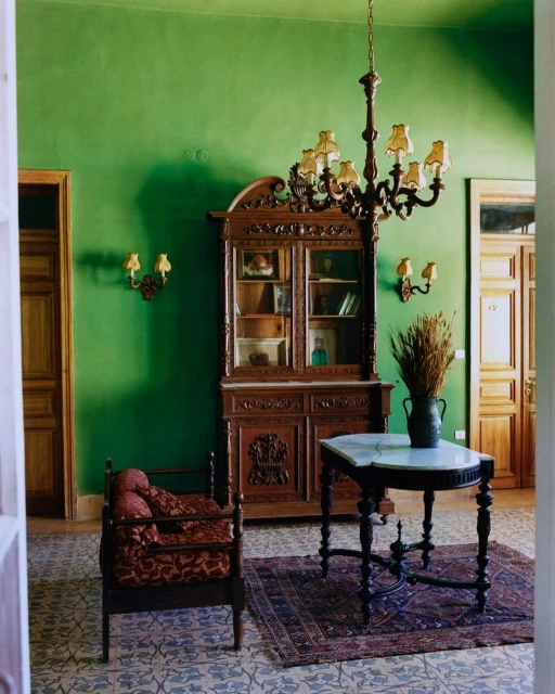 Interior design featuring an antique wooden cabinet, ornate chandelier, small table with a marble top, and patterned rug in a green room.