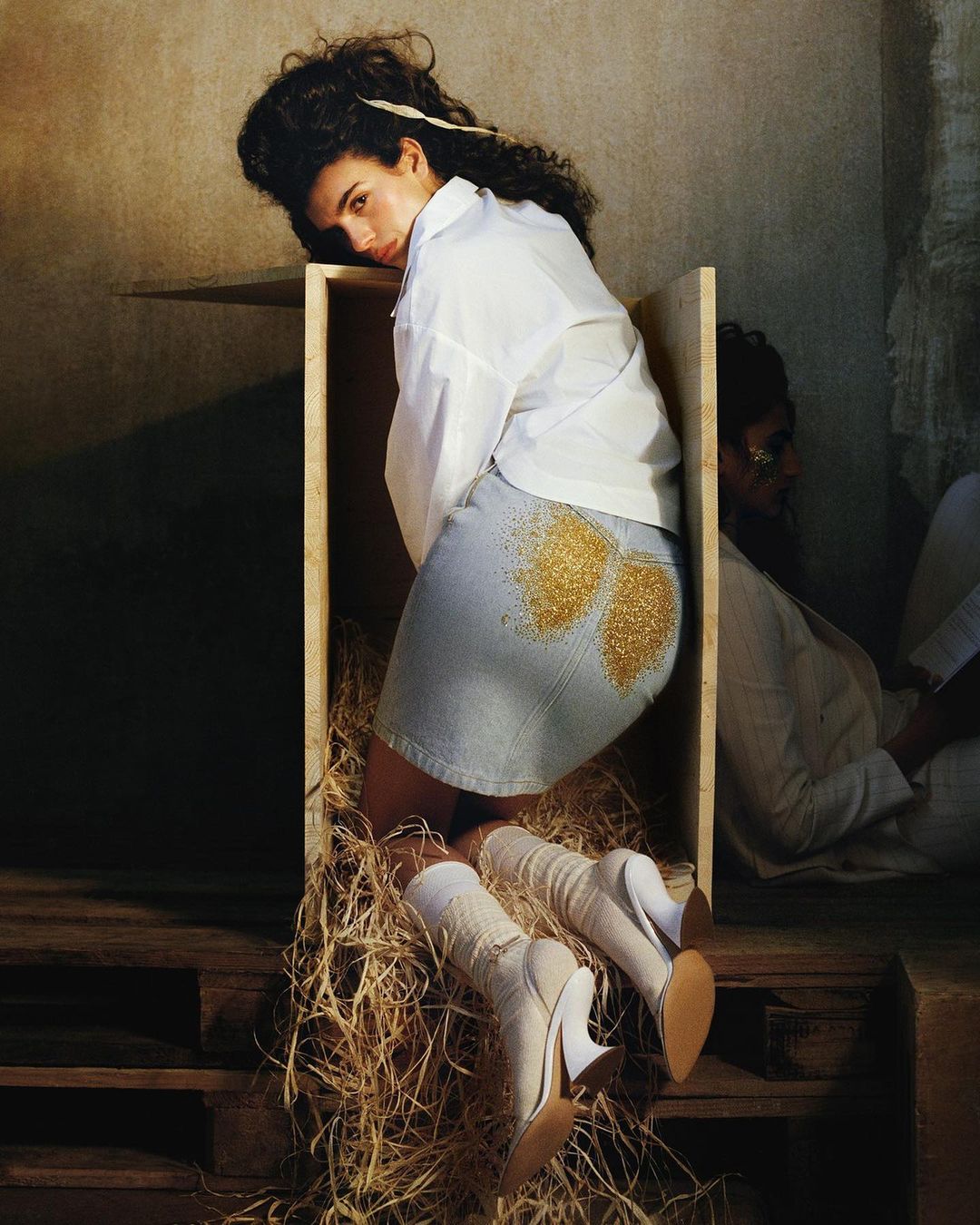 Portrait of a woman with curly hair wearing a white shirt and denim skirt with gold glitter, posing in a wood box on a straw-covered surface.