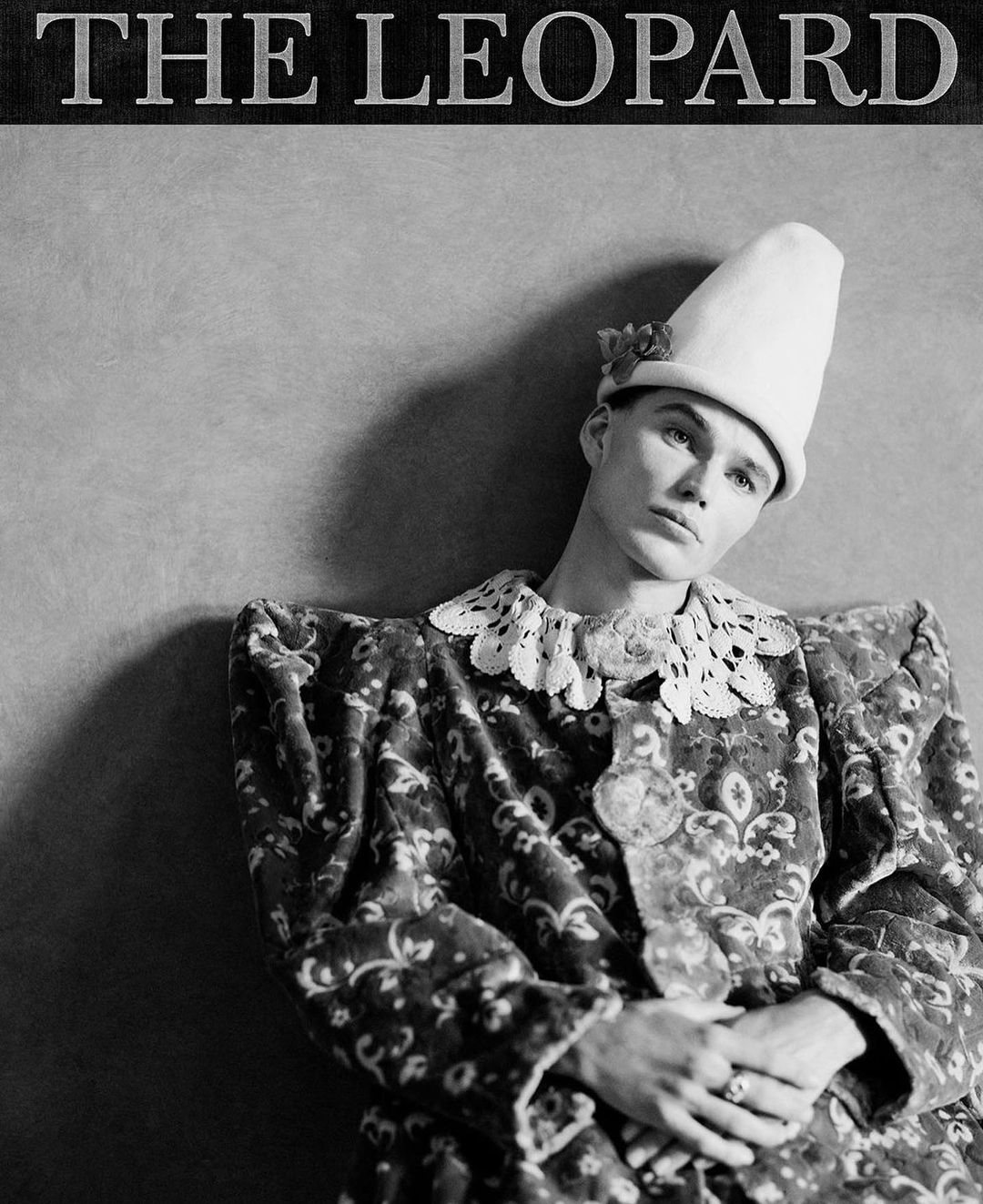 Black and white portrait of person in patterned jacket, lace collar, cone hat with flower, and ring in front of a grey wall.