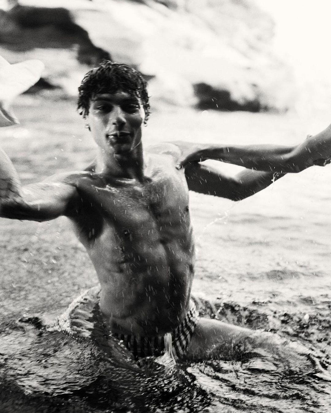 Black and white medium shot of a muscular adult male emerging from water, showcasing his physique and interaction with the water.