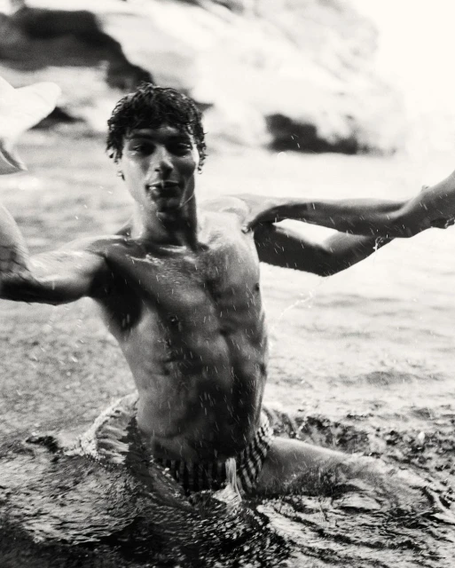 Black and white medium shot of a muscular adult male emerging from water, showcasing his physique and interaction with the water.