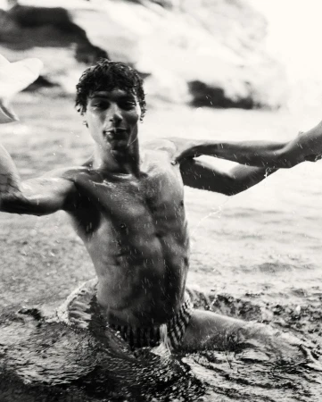 Black and white medium shot of a muscular adult male emerging from water, showcasing his physique and interaction with the water.