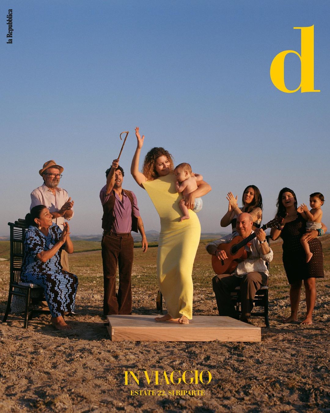 Editorial shot of a woman dancing on a wooden platform with a baby in her arms, surrounded by a group of people in a field.