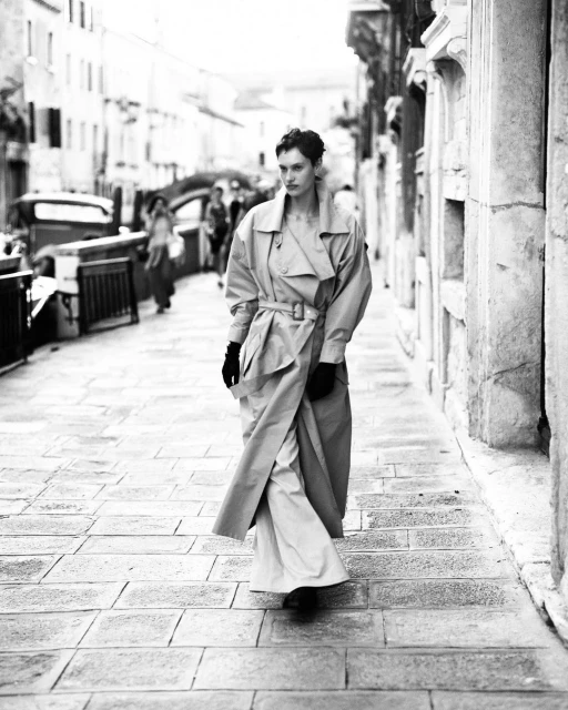 Woman walks down a cobblestone street wearing a long trench coat and pants. Buildings line the street in the background in black and white.