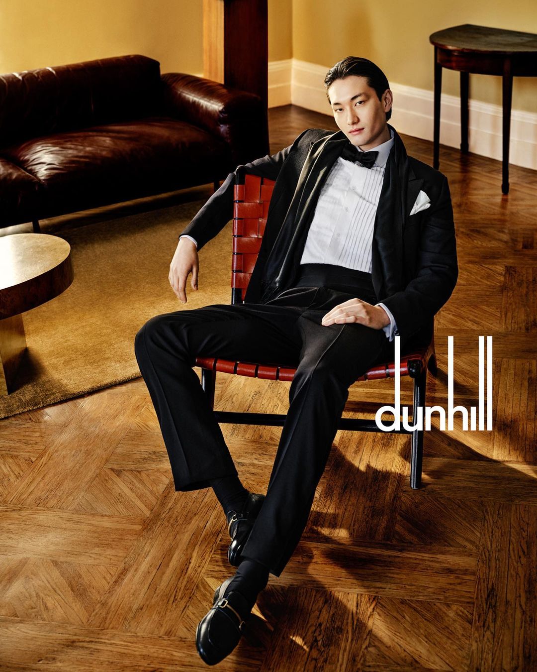 Asian man in black tuxedo seated in a leather chair inside an elegant room with hardwood floors, photographed by Brett Lloyd.