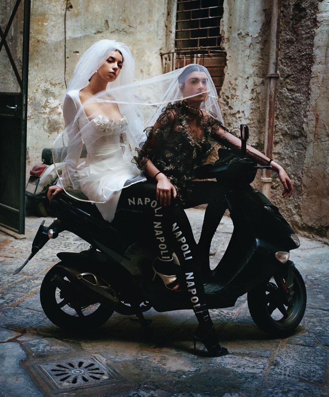 Two women in wedding dresses pose on a moped in an urban alleyway with weathered stone walls, creating a fashion editorial shot.