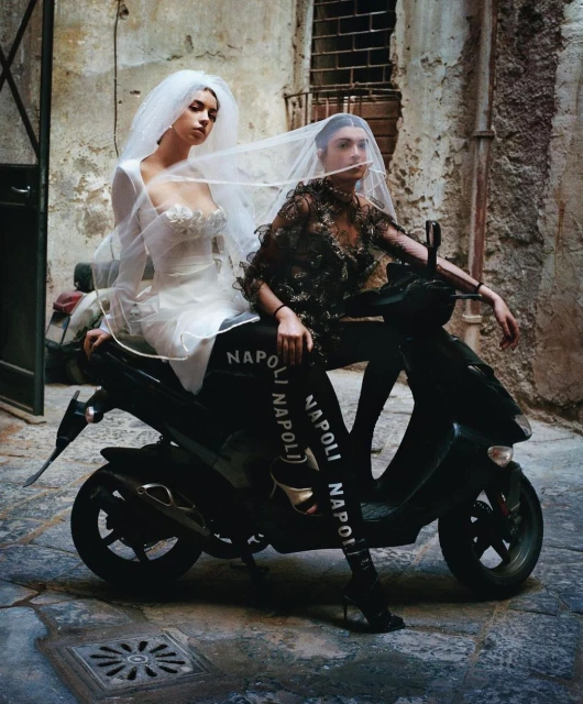 Two women in wedding dresses pose on a moped in an urban alleyway with weathered stone walls, creating a fashion editorial shot.