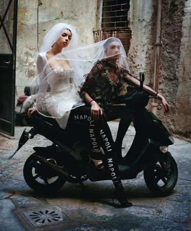 Two women in wedding dresses pose on a moped in an urban alleyway with weathered stone walls, creating a fashion editorial shot.