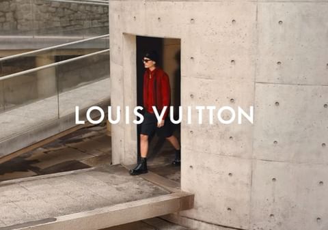 A person in a red sweater and black shorts exits a concrete doorway in an outdoor setting with the Louis Vuitton logo overlayed.