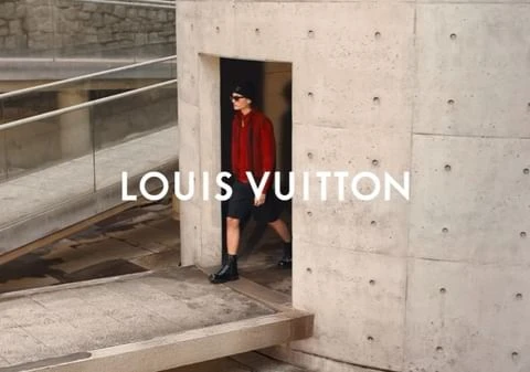 A person in a red sweater and black shorts exits a concrete doorway in an outdoor setting with the Louis Vuitton logo overlayed.
