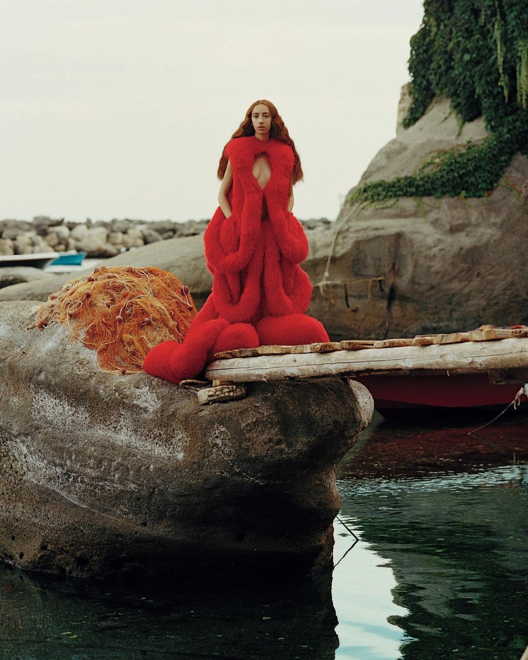Fashionable woman in oversized red fuzzy outfit stands on rocks by the sea, boat and green foliage background under cloudy sky.