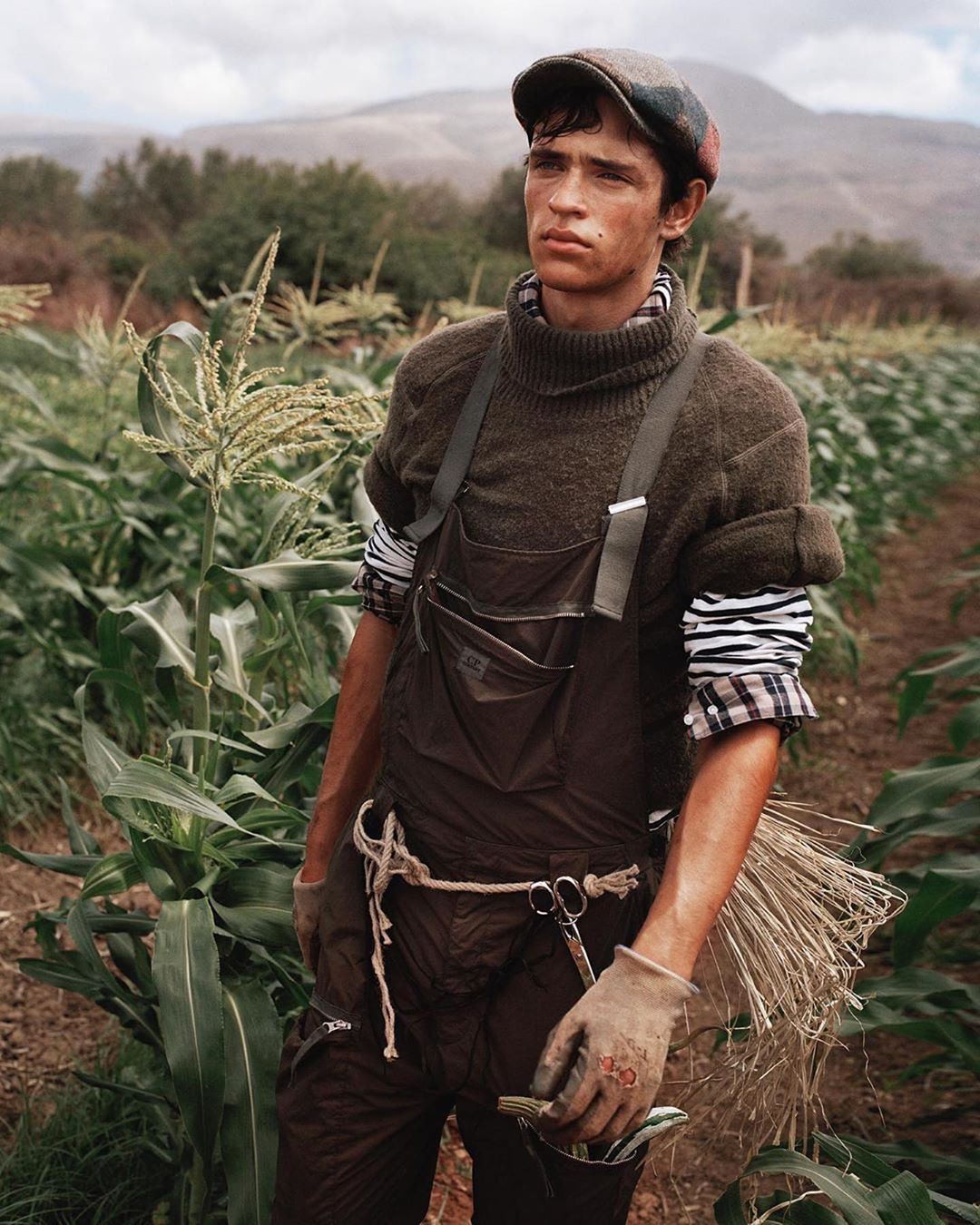 Male model in cornfield wearing overalls and cap. Outdoor setting with natural light, conveying a rural and agricultural atmosphere.