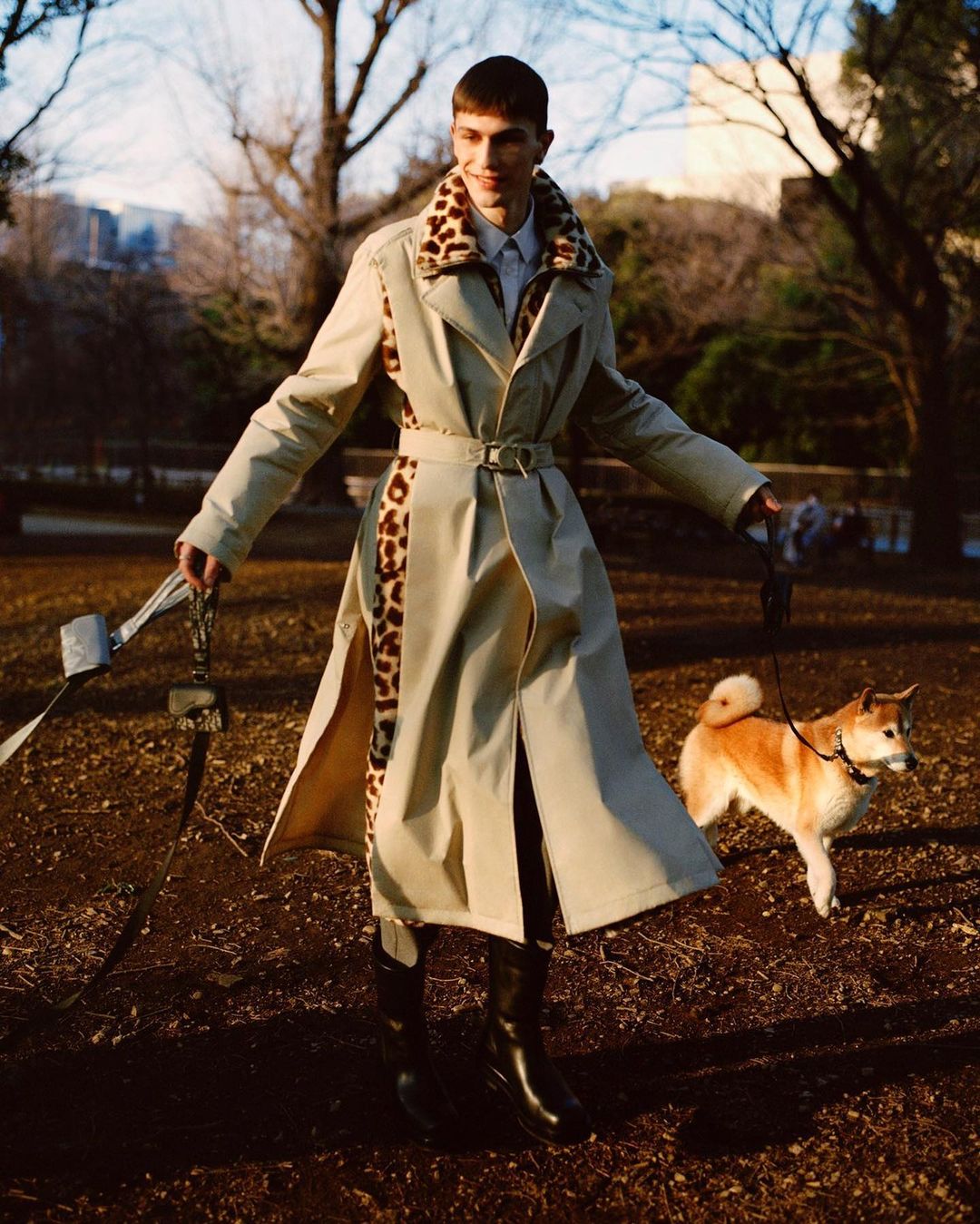 Male fashion model in trench coat walks tan Shiba Inu dog on leash through park in editorial fashion shoot on overcast day.