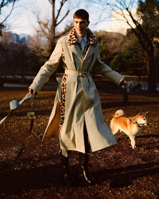 Male fashion model in trench coat walks tan Shiba Inu dog on leash through park in editorial fashion shoot on overcast day.