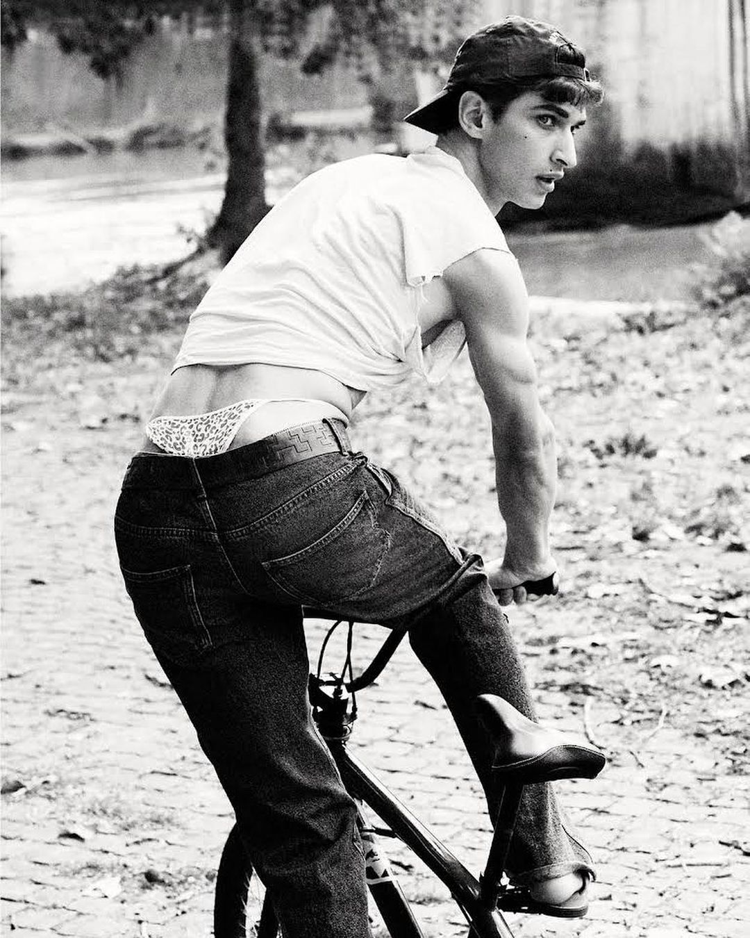 Black and white shot of a young adult man riding a bicycle on a cobblestone road, wearing jeans and a backward baseball cap outdoors.