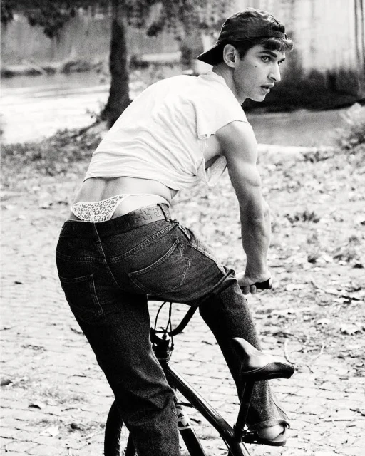 Black and white shot of a young adult man riding a bicycle on a cobblestone road, wearing jeans and a backward baseball cap outdoors.