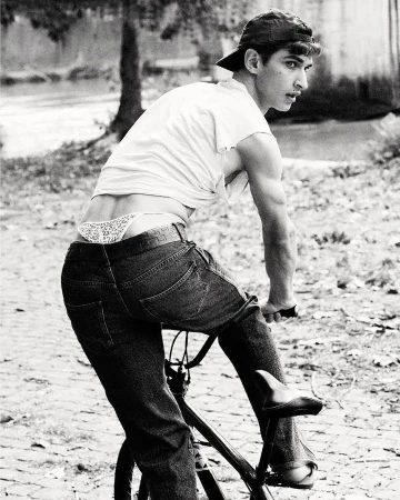 Black and white shot of a young adult man riding a bicycle on a cobblestone road, wearing jeans and a backward baseball cap outdoors.