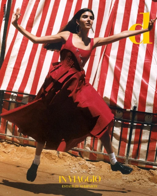 A woman in a red dress is joyfully leaping in the air, arms outstretched, with a red and white striped awning in the background.