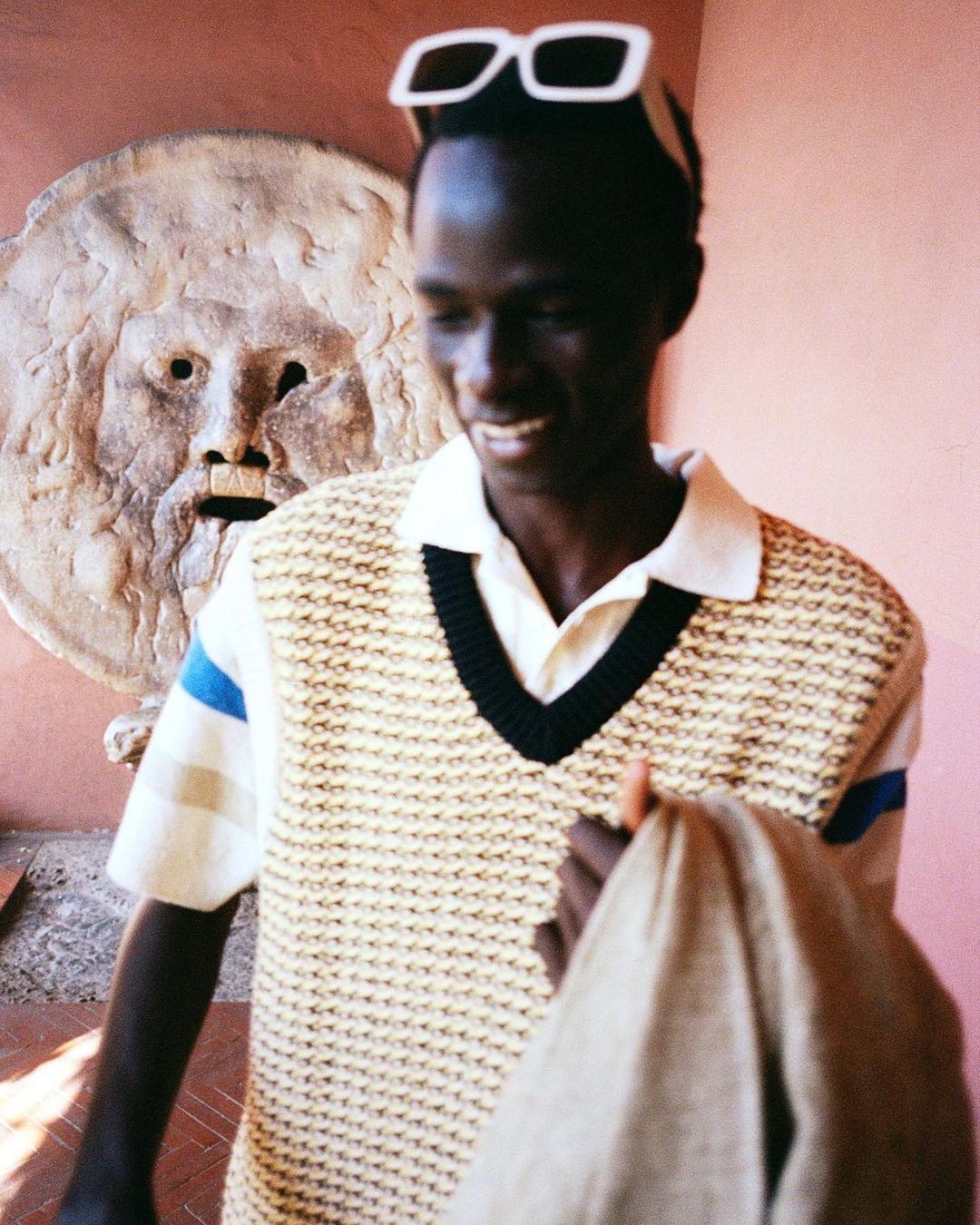 Smiling adult male wearing sunglasses and knit vest poses in front of a stone mask carving set against a pale pink wall, medium shot.