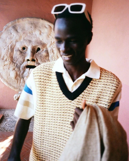 Smiling adult male wearing sunglasses and knit vest poses in front of a stone mask carving set against a pale pink wall, medium shot.