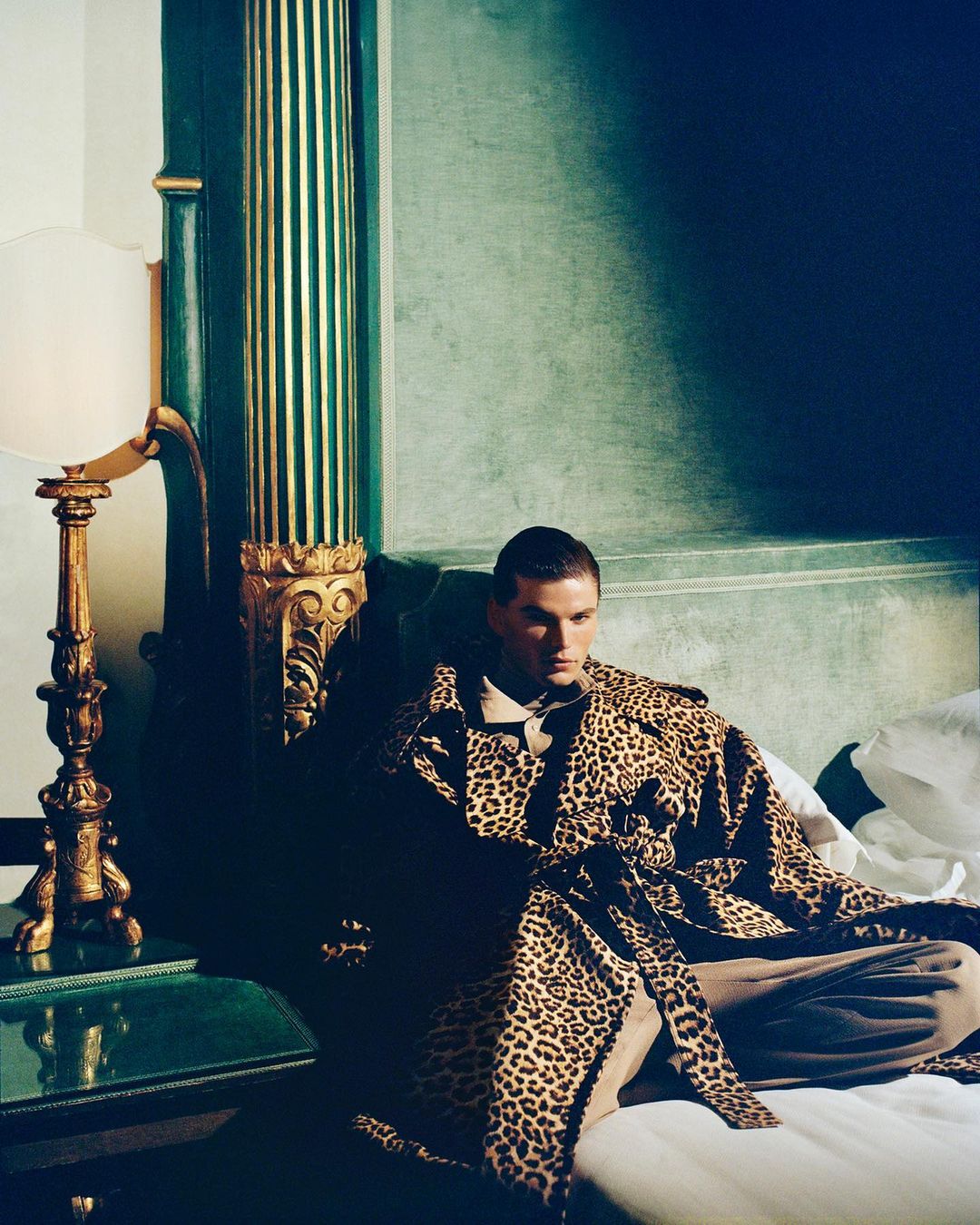 Stylish adult male model in a leopard print coat lounges in a hotel room with vintage decor and soft lighting, showcasing fashion and lifestyle.
