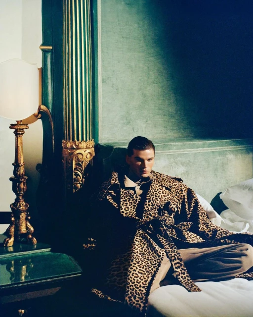 Stylish adult male model in a leopard print coat lounges in a hotel room with vintage decor and soft lighting, showcasing fashion and lifestyle.