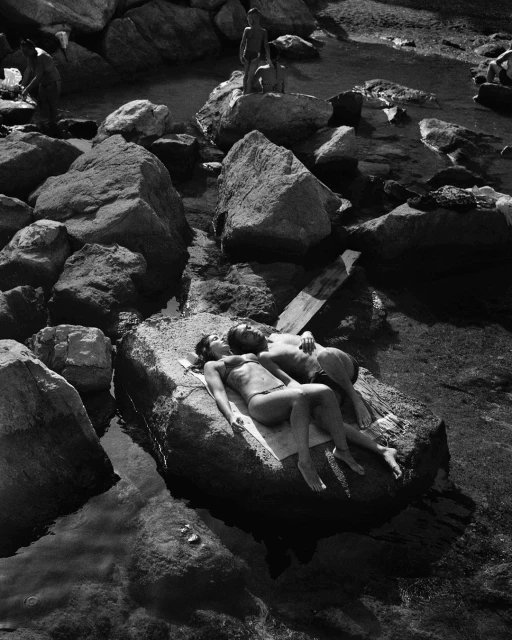 Monochrome shot of a couple sunbathing on a rock near water, with other people on rocks in the background, creating a stark, vintage-style image.