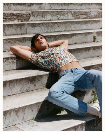 Young adult male poses on stone steps wearing patterned top and jeans, bathed in natural light outdoors in a fashion setting.