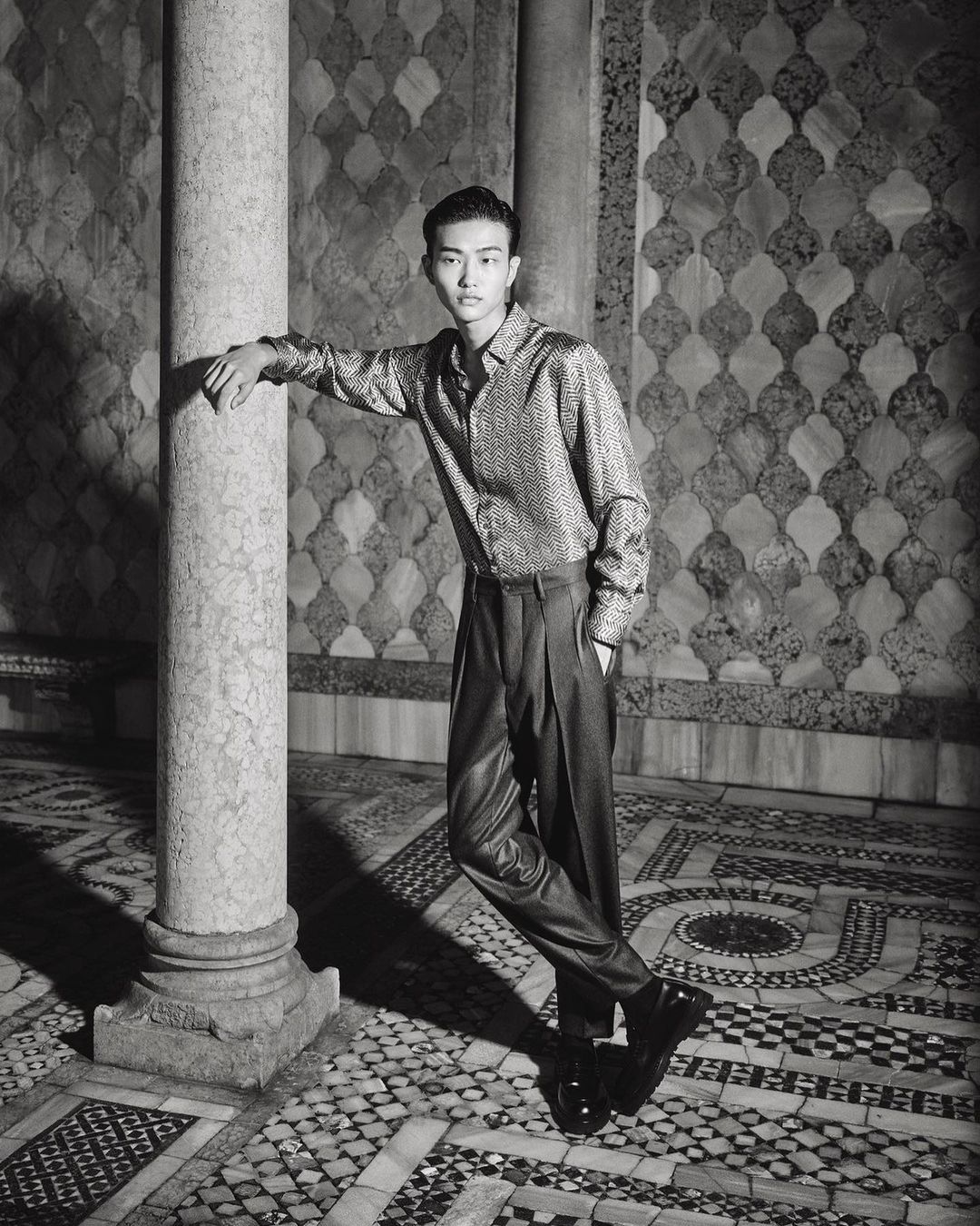 Young Asian man in shiny shirt and dark pants poses by a marble pillar against an ornate tiled wall, fashion photography.