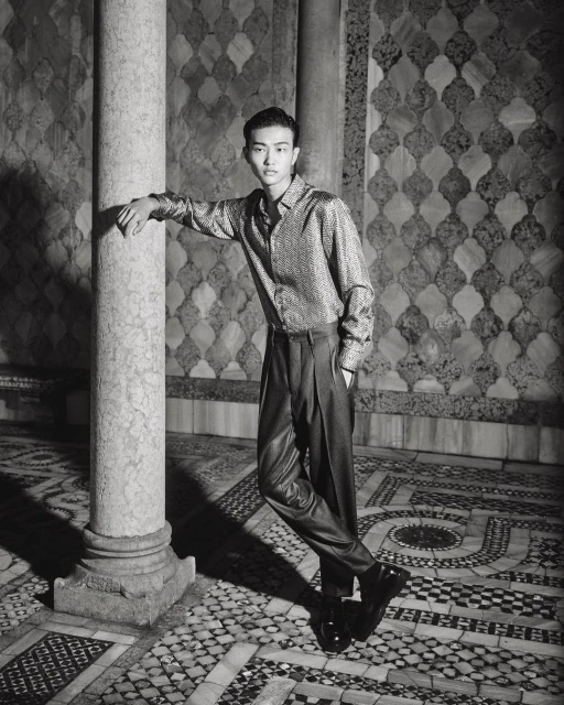 Young Asian man in shiny shirt and dark pants poses by a marble pillar against an ornate tiled wall, fashion photography.