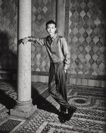 Young Asian man in shiny shirt and dark pants poses by a marble pillar against an ornate tiled wall, fashion photography.