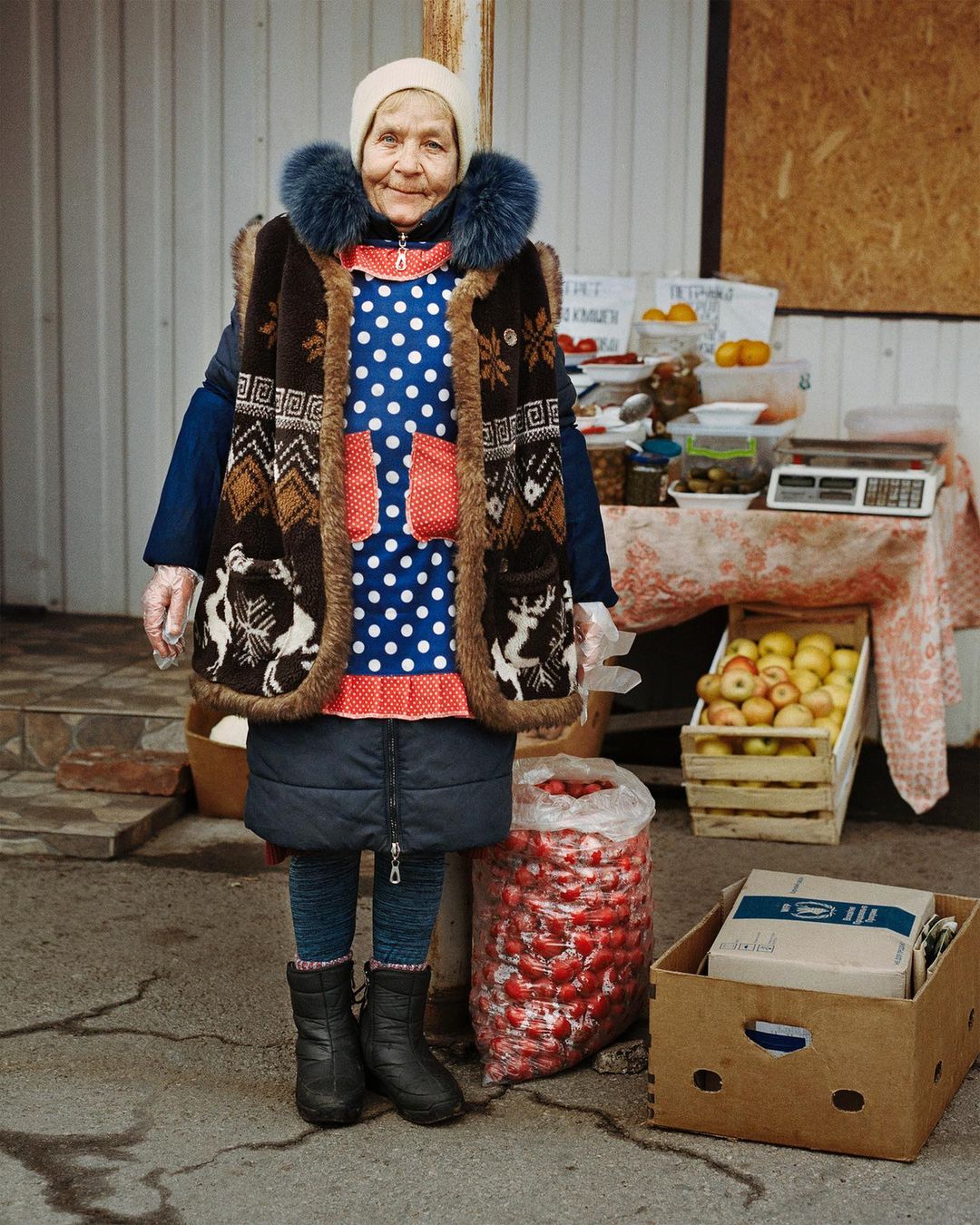 Elderly woman at outdoor market stands by fruit and vegetables, wearing a polka dot dress, vest, and winter boots on a sunny day.