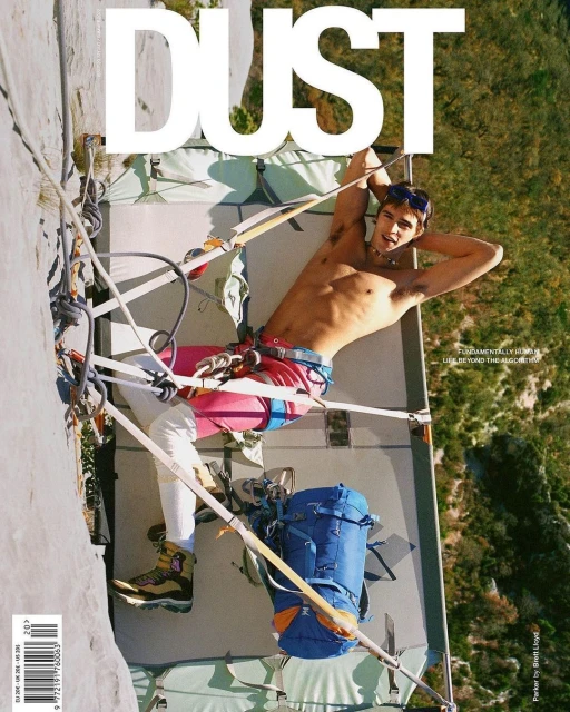 Shirtless man lays on a portaledge fixed to a rock cliff face with a backpack. A magazine cover with the word DUST is visible.